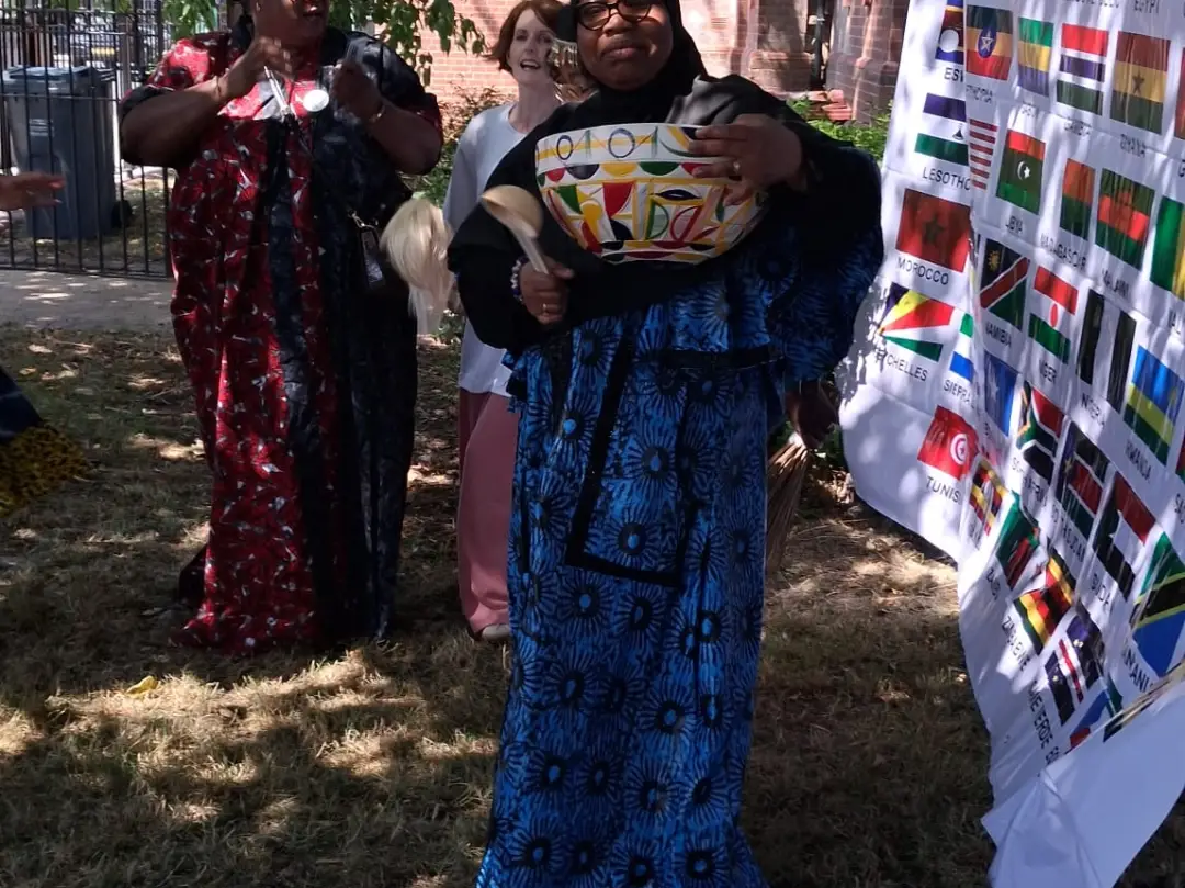 A woman in blue patterned dress holding a decorated bowl outdoors, beside a large banner printed with the flags of African nations.